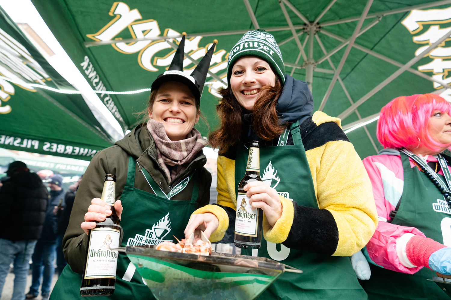 Zwei Frauen mit Kopfbedeckung und Bierflasche in der Hand unter einem grünen Sonnenschirm