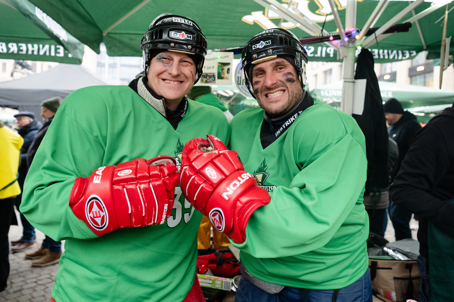 Zwei Männer in grünem Trikot,  Eishockey-Helm und roten Eishockey-Handschuhen