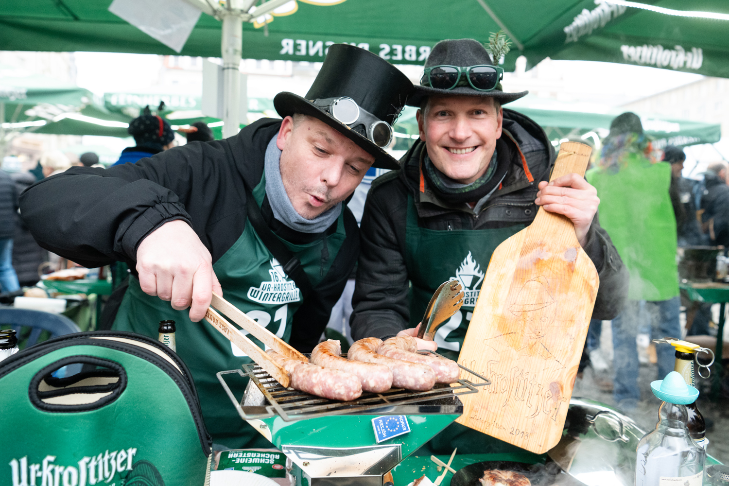 Zwei Männer mit Kopfbedeckung vor einem Grill mit Würsten