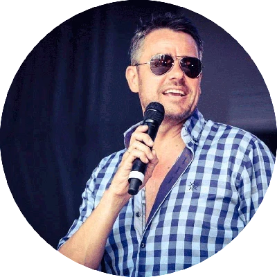 Ein Mann in einem blau-weiß karierten Hemd hält ein Mikrofon und lächelt.