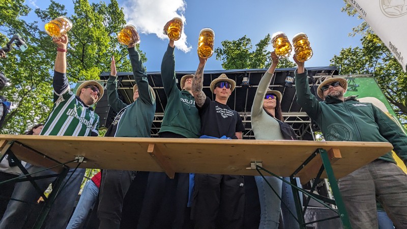 6 Personen stehen hinter einem Biertisch und halten gefüllte Biergläser hoch.