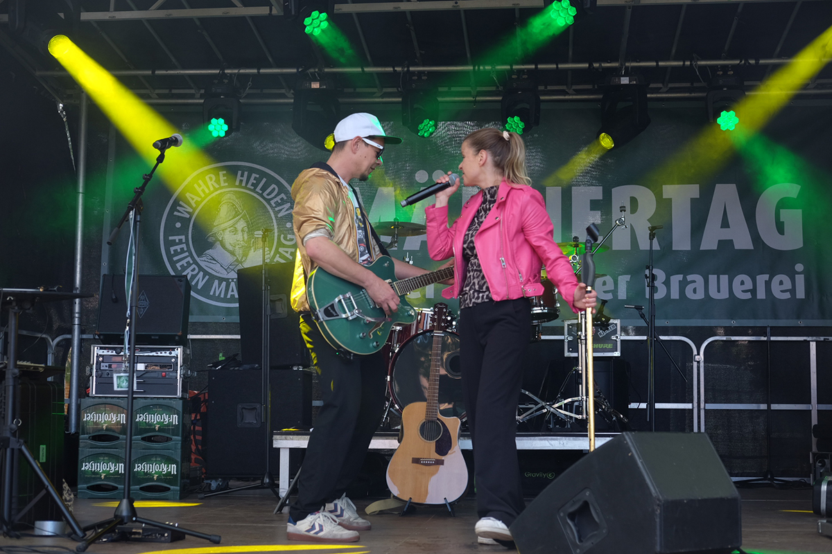Ein Gitarrist und eine Sängerin performen auf einer Bühne, im Hintergrund bunte Lichter.