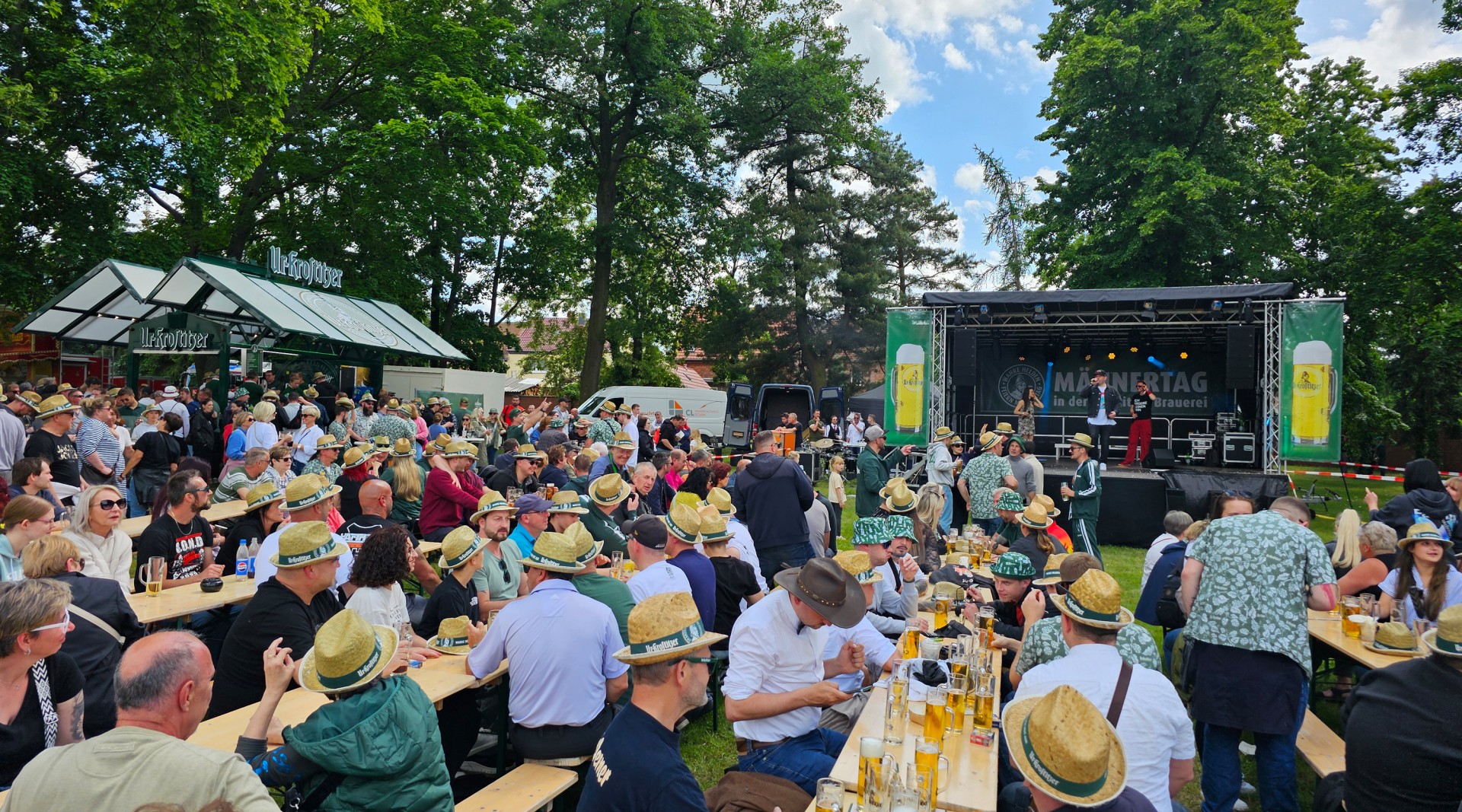 Viele Menschen sitzen auf Biergarnituren vor einer Bühne und feiern den  Männertag, im Hintergrund befinden sich Bäume.