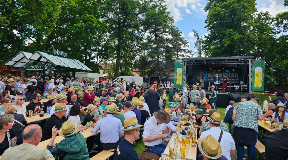 Viele Menschen sitzen auf Biergarnituren vor einer Bühne und feiern den  Männertag, im Hintergrund befinden sich Bäume.