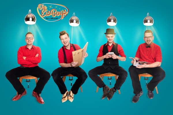 Vier rot schwarz gekleidete Männer sitzen nebeneinanderauf Stühlen, jeder unter einer Lampe.