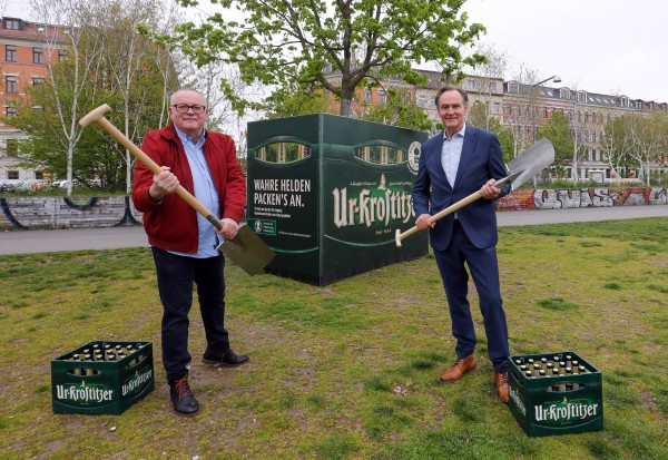 Zwei Männer stehen in einem Park mit Schaufeln neben einer großen grünen Box und Bierkisten.