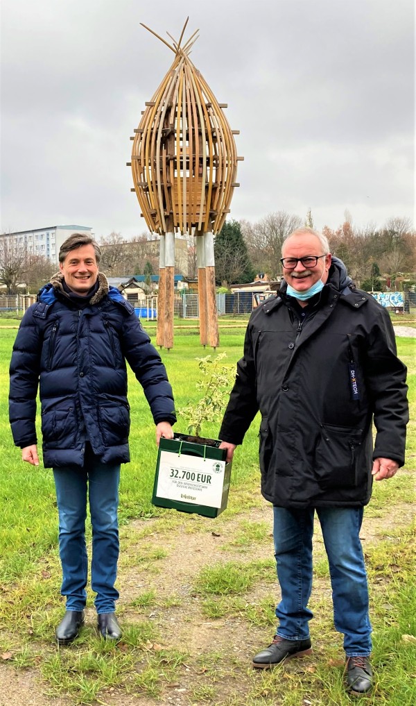 Zwei Männer stehen lächelnd vor einer großen hölzernen Skulptur und halten eine Spendentafel.