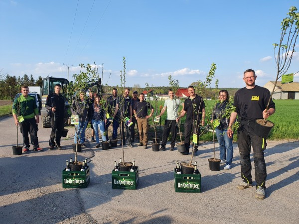 Elf Personen stehen lächelnd auf einer Straße und halten jeweils einen kleinen Baum. Sie tragen Freizeitkleidung und Arbeitskleidung.