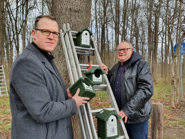 Zwei Männer montieren Vogelhäuser an einem Baum nahe einem Waldweg.