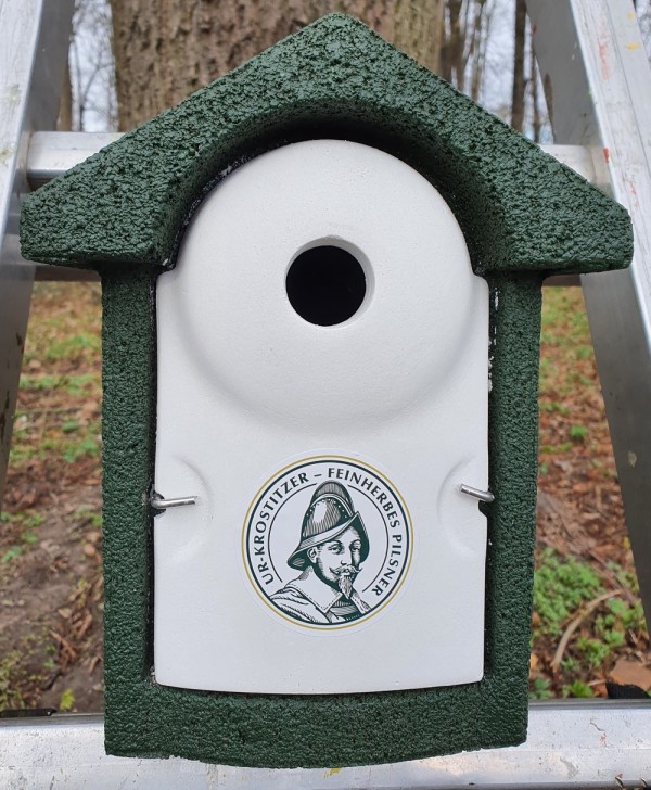 Ein Vogelhaus aus weißen und grünen Flächen ist an einem Metallständer befestigt. Es hat eine runde Öffnung und eine illustrierte Plakette.