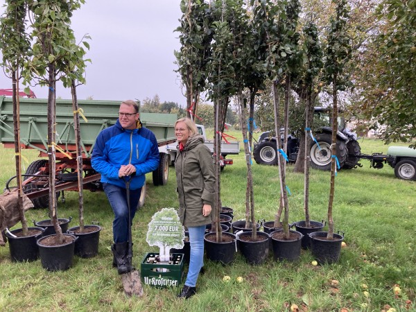 Ein Mann und eine Frau stehen neben jungen Bäumen auf einer Wiese. Sie halten Spaten und lächeln neben einem Schild mit 
