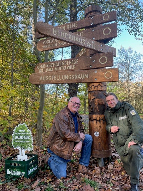 Zwei Männer stehen lächelnd neben einem Wegweiser aus Holz im Wald.