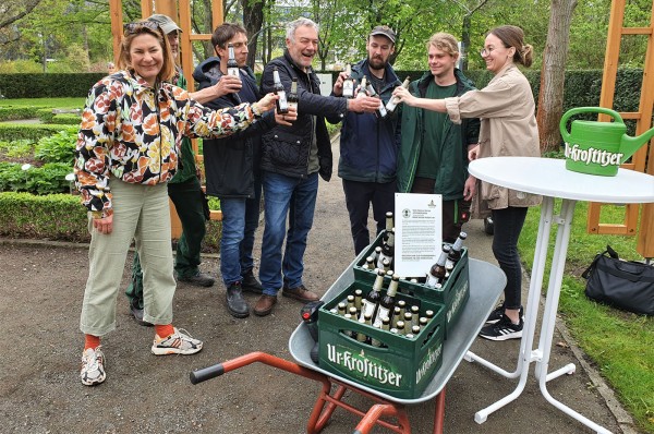 Eine Gruppe Menschen stößt im Freien mit Bierflaschen an, neben ihnen steht eine Schubkarre mit weiteren Flaschen.