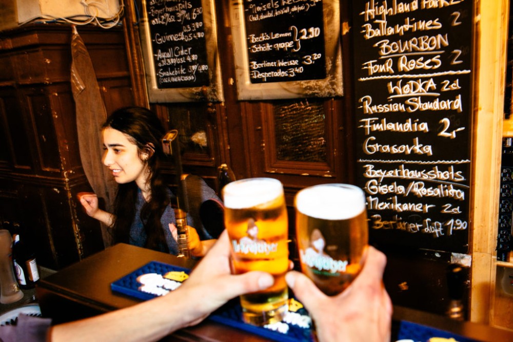 Eine Person reicht in einer Bar zwei Biergläser über den Tresen. Im Hintergrund sind Getränkepreise auf einer Tafel sichtbar.