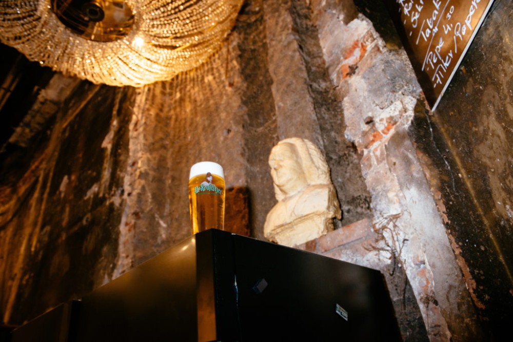 Ein Bierglas steht auf einem Tresen im Vordergrund, dahinter eine Büste neben rauer Wand.