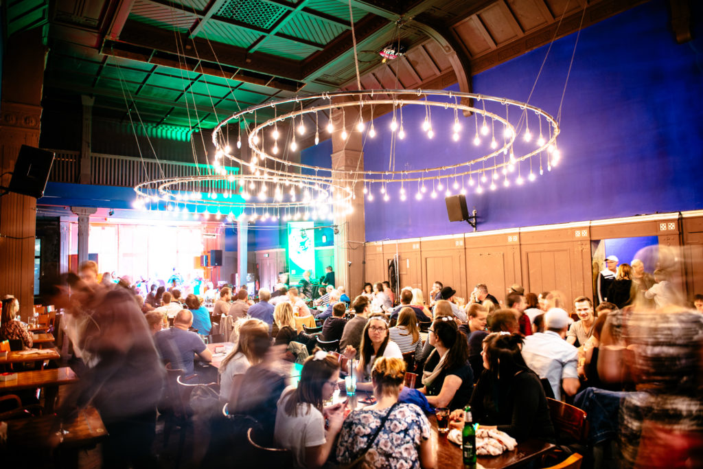 Viele Menschen sitzen in einem beleuchteten Saal und schauen auf eine Bühne.