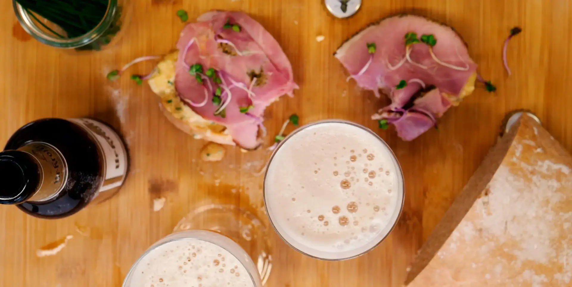 Zwei Gläser Bier stehen neben einem Teller mit belegten Broten auf einem Holztisch.