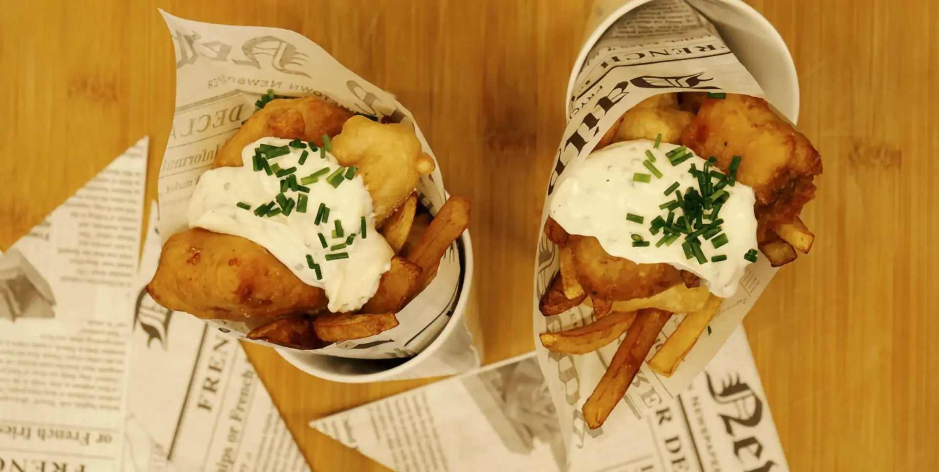 Zwei Portionen Fish and Chips in kegelförmigen Behältern stehen auf einem Holztisch.