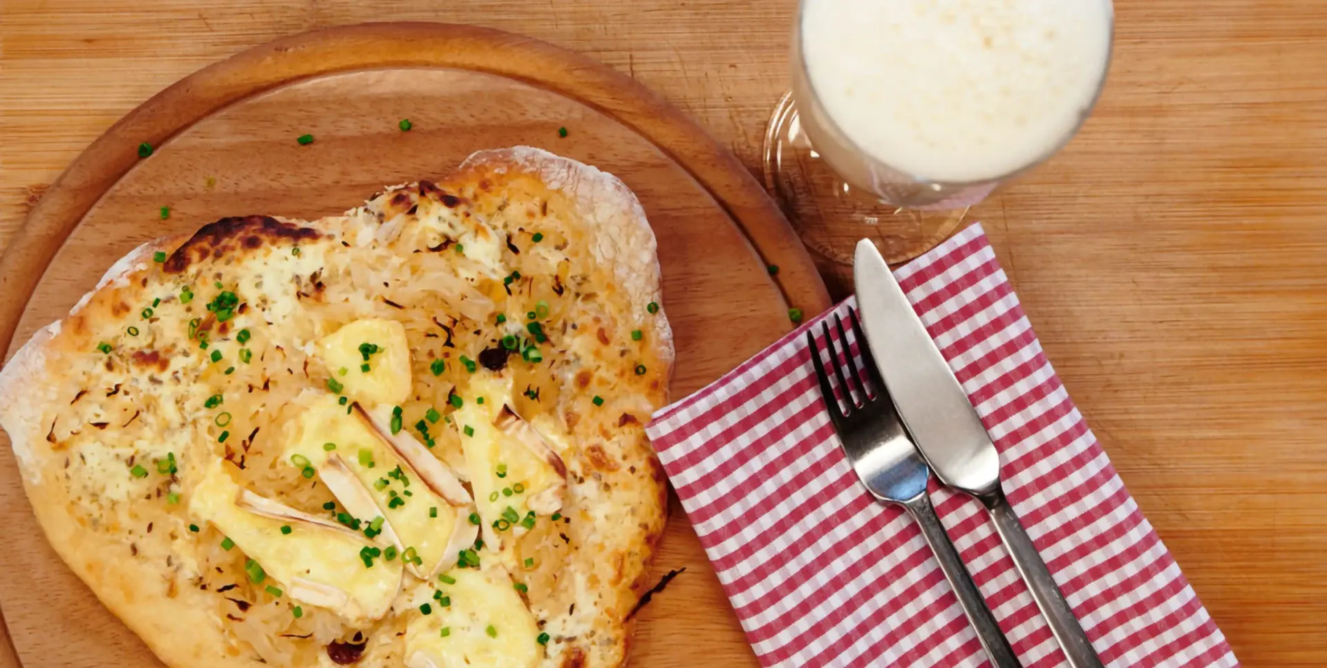 Eine frisch gebackene Pizza mit Käse und Schnittlauch liegt auf einem Holzbrett, daneben ein Glas Bier und Besteck auf einem rot-weiß karierten Tuch.