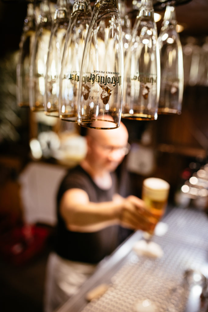 Ein Barkeeper serviert ein Bierglas, im Hintergrund hängen unscharfe Biergläser.