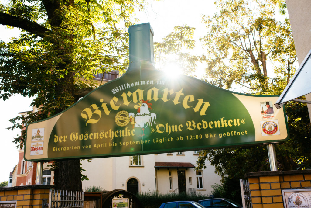 Grünes Werbeschild eines Biergartens zeigt Öffnungszeiten und illustrierte Bestandteile.