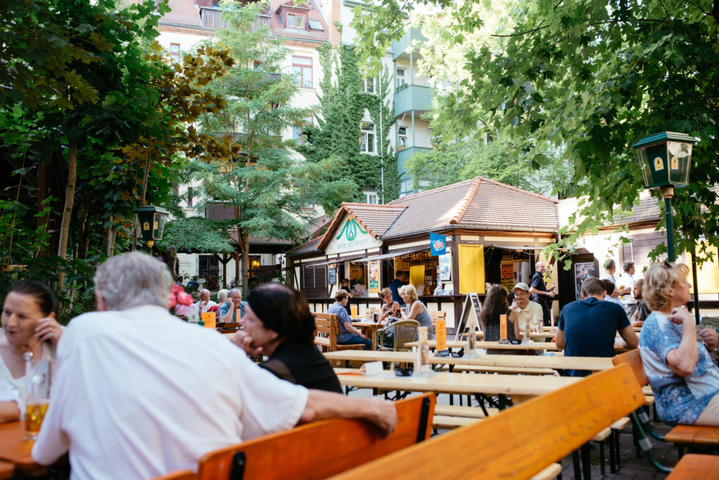 Menschen sitzen im Freien in einem belebten Biergarten umgeben von Bäumen und Gebäuden.