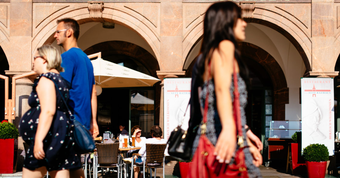 Mehrere Personen gehen an einem sonnigen Tag vor einem Café mit Außenbestuhlung entlang.