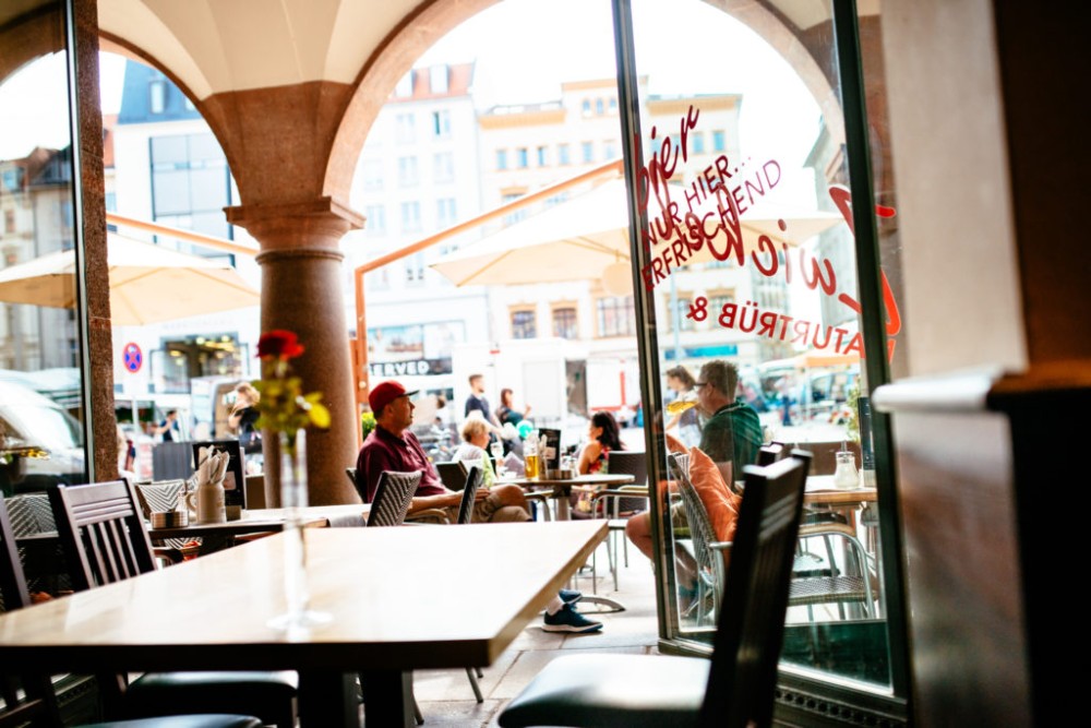 Eine Caféterrasse unter Arkaden mit Gästen, die Getränke genießen, und Blick auf eine belebte Straße.