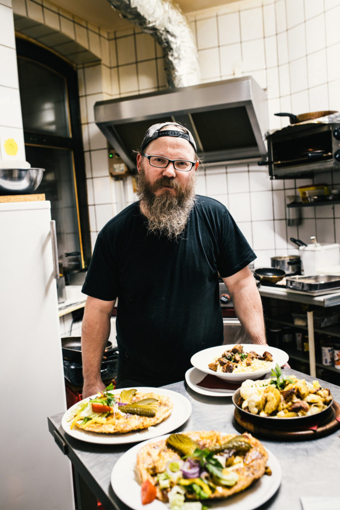 Ein Koch mit Bart steht in einer Küche und hält Teller mit Essen.