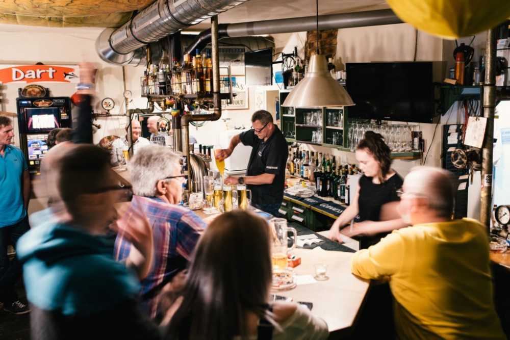 Mehrere Personen unterhalten sich in einer belebten Bar, während zwei Barkeeper Getränke servieren.
