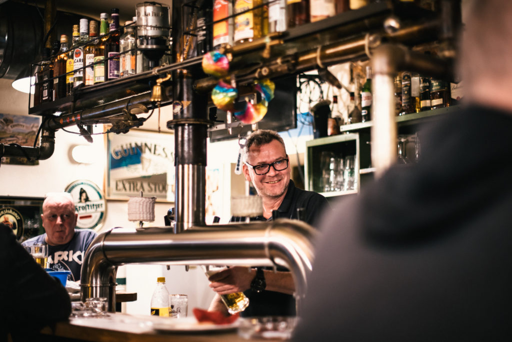 Ein lachender Mann bedient hinter einer Bar, im Hintergrund sitzt ein älterer Herr.
