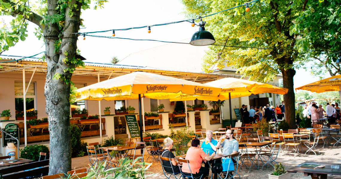 Menschen sitzen an Tischen unter gelben Sonnenschirmen in einem Outdoor-Café.