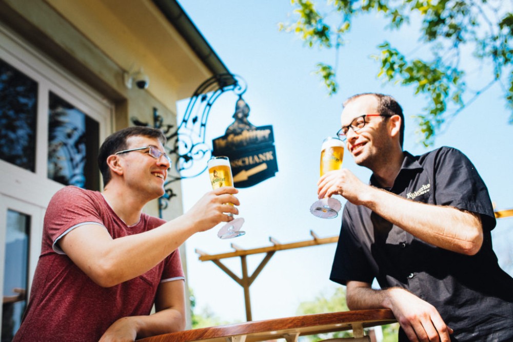 Zwei Männer sitzen lachend im Freien und stoßen mit Biergläsern an.