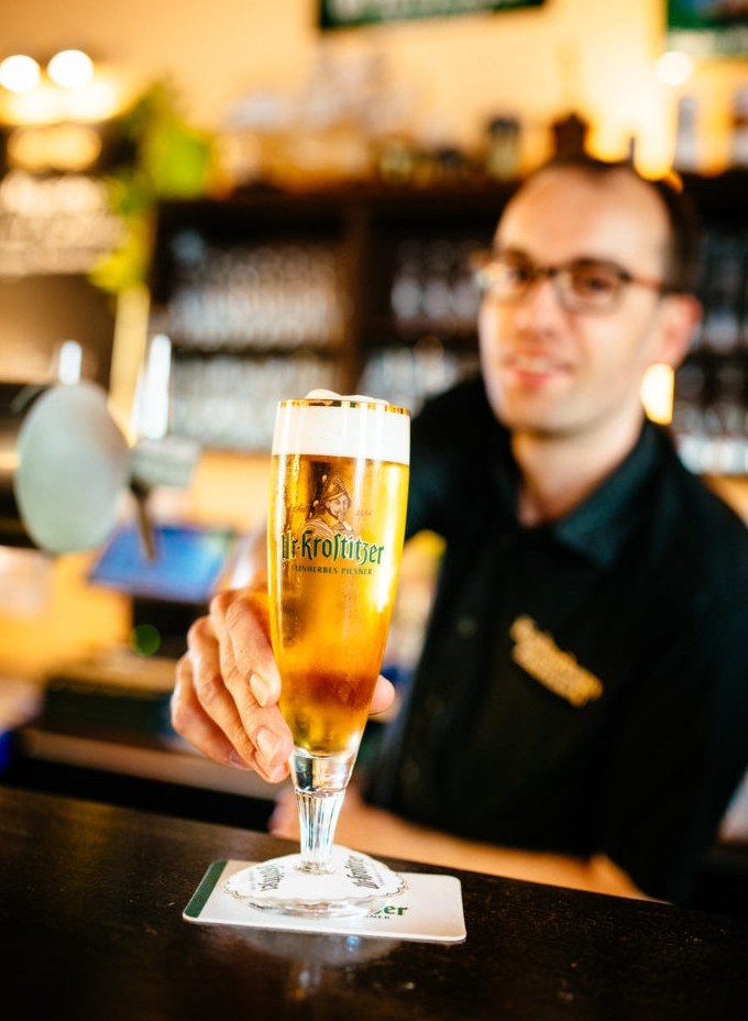 Ein Mann in dunkler Kleidung reicht ein gefülltes Bierglas über eine Bartheke.
