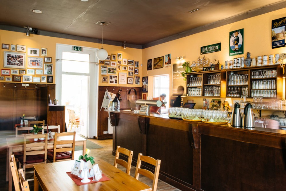 Innenansicht eines gemütlichen Cafés mit Holzmöbeln und einer Bartheke, an den Wänden hängen zahlreiche Fotos.