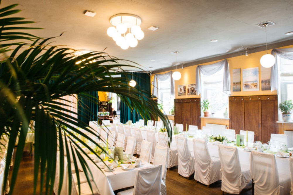 Ein festlich gedeckter Speisesaal mit weißen Stuhlbezügen und Tischdecken. Grüne Pflanzen schmücken den Raum, ergänzt durch natürliche Beleuchtung.