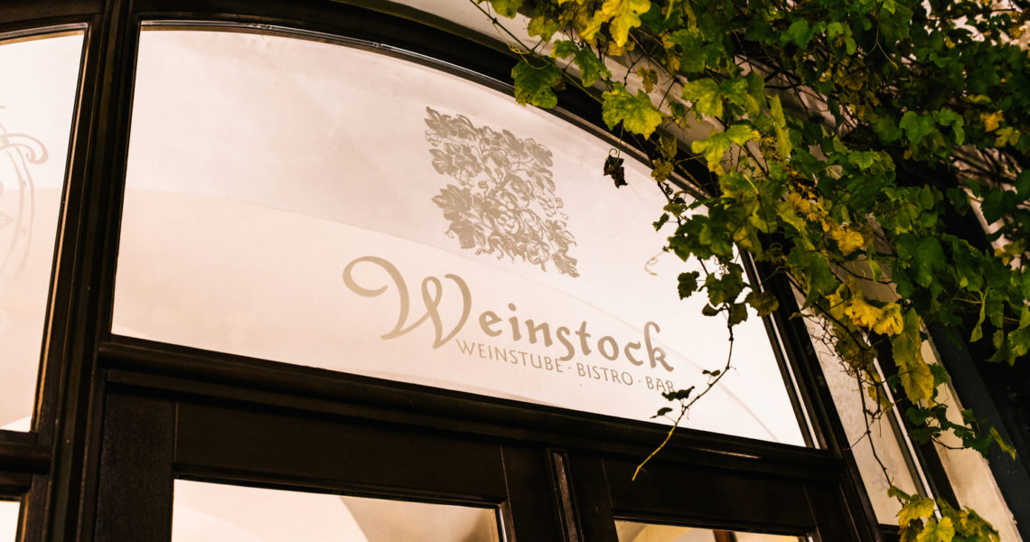 Ein Fenster eines Gebäudes mit dekorativer Beschriftung und darüber rankenden grünen Pflanzen.