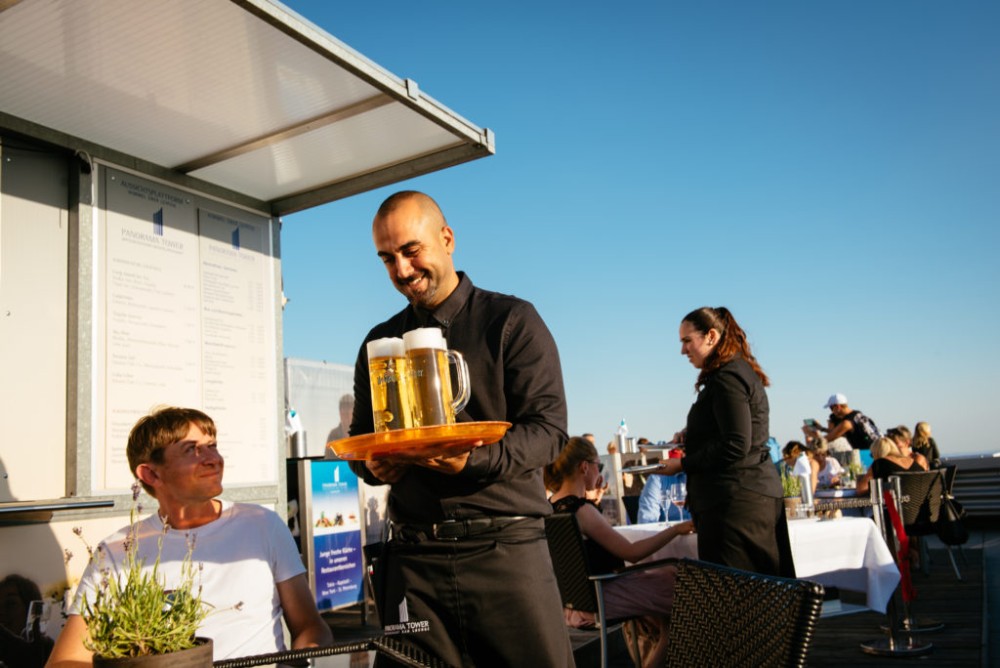 Ein Kellner serviert einem lächelnden Gast Biere auf einer Dachterrasse im Freien.