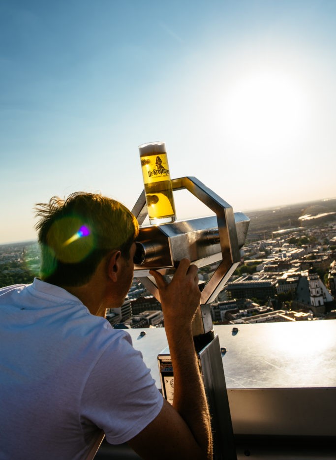 Eine Person betrachtet die Stadtsilhouette durch ein Fernrohr, neben dem ein Glas Bier steht.