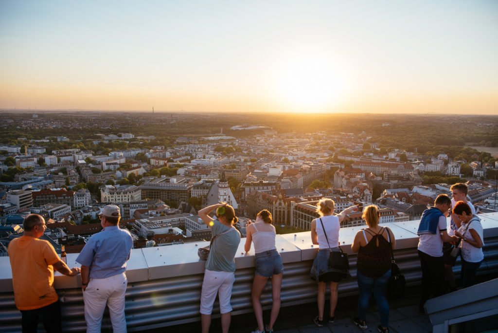 Menschen betrachten vom Dach eines hohen Gebäudes den Sonnenuntergang über einer Stadt.