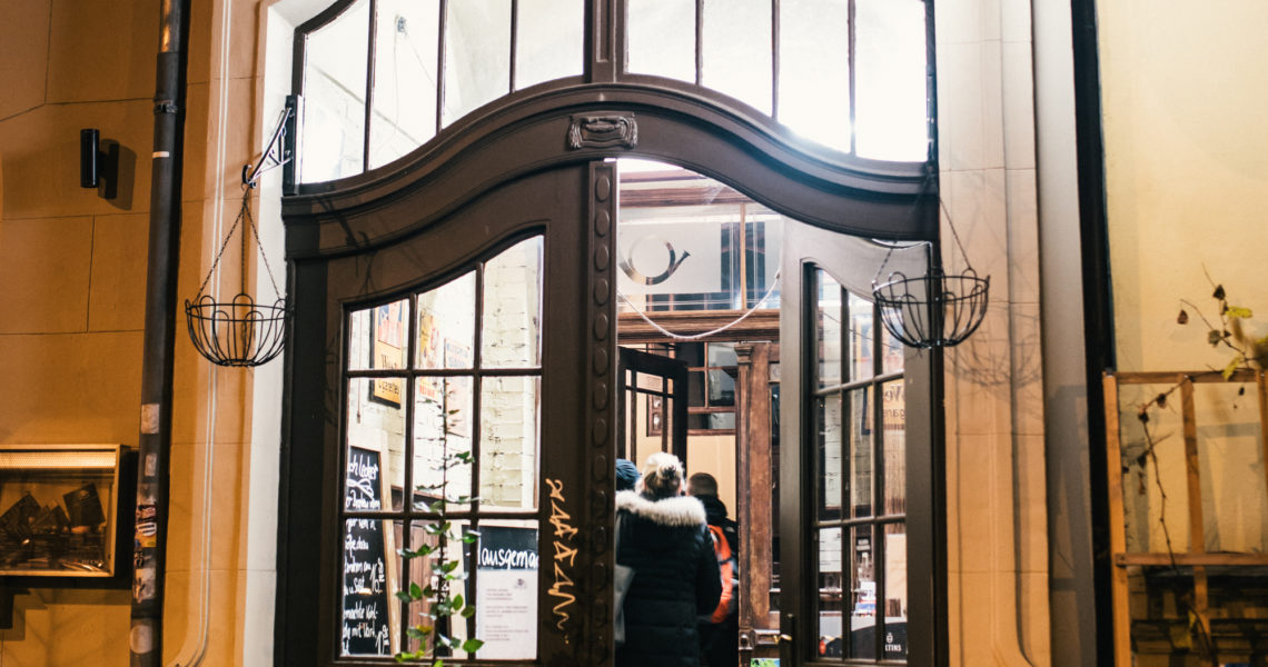 Zwei Personen stehen vor einer offenen, gebogenen Tür eines stilvollen Cafés.
