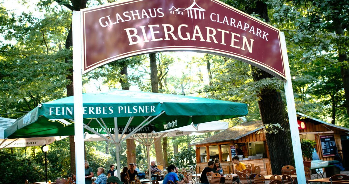Besucher genießen Getränke unter grünen Sonnenschirmen in einem Biergarten mit Schild.