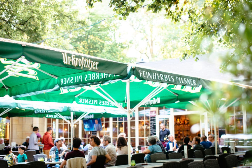 Menschen sitzen entspannt unter grünen Sonnenschirmen in einem Außencafé.