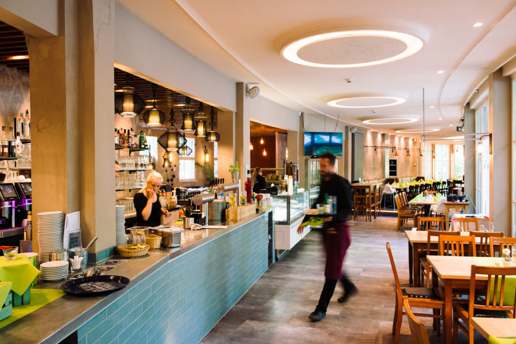 Ein modernes Restaurant mit belebter Bar und Bedienung in Bewegung. Lichter hängen über dem Tresen und Tische sind verteilt.