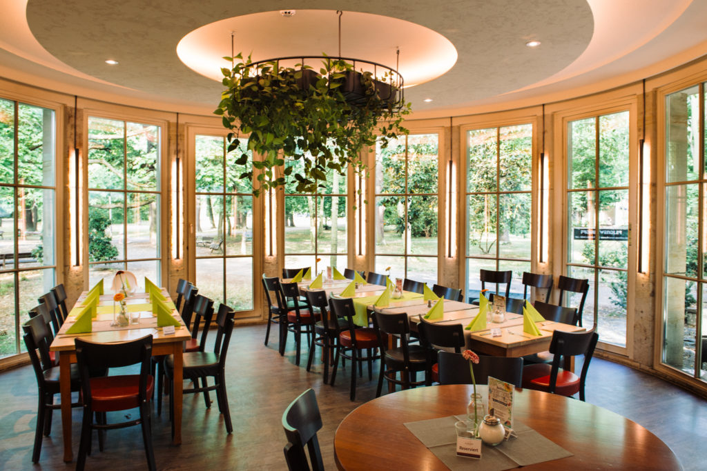 Ein heller Speiseraum mit runden und rechteckigen Tischen, umgeben von großen Fenstern.