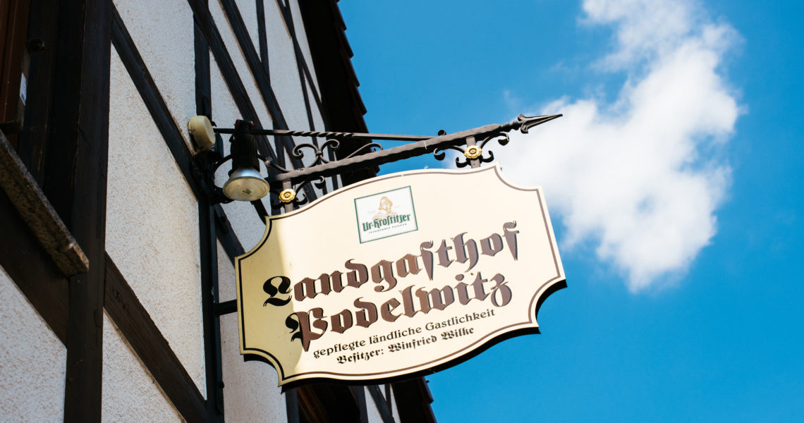 Schild eines Landgasthofs hängt an einem schwarzen, geschwungenen Metallhalter vor einem Fachwerkhaus.