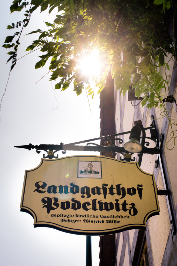 Ein Schild des Landgasthofs Bodelwitz hängt vor einem mit Blättern bedeckten Ast. Die Sonne scheint hell durch die Blätter.