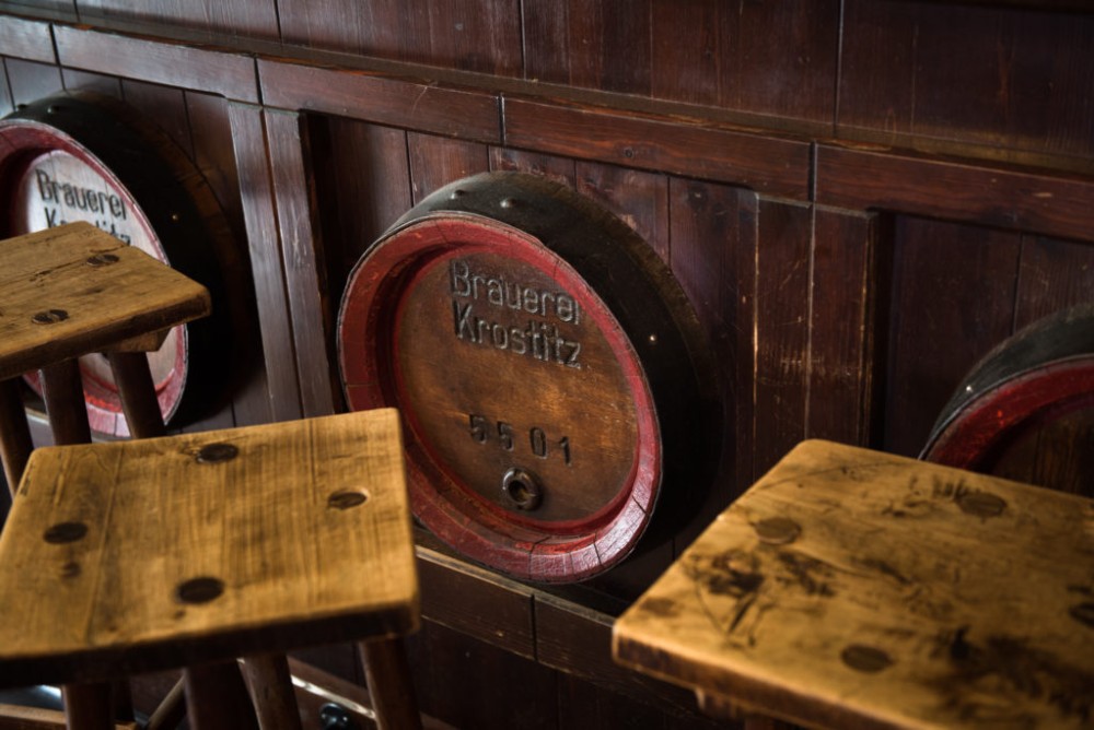 Drei Holzfässer in einer Reihe an einer dunklen Wand und hölzerne Tische davor.
