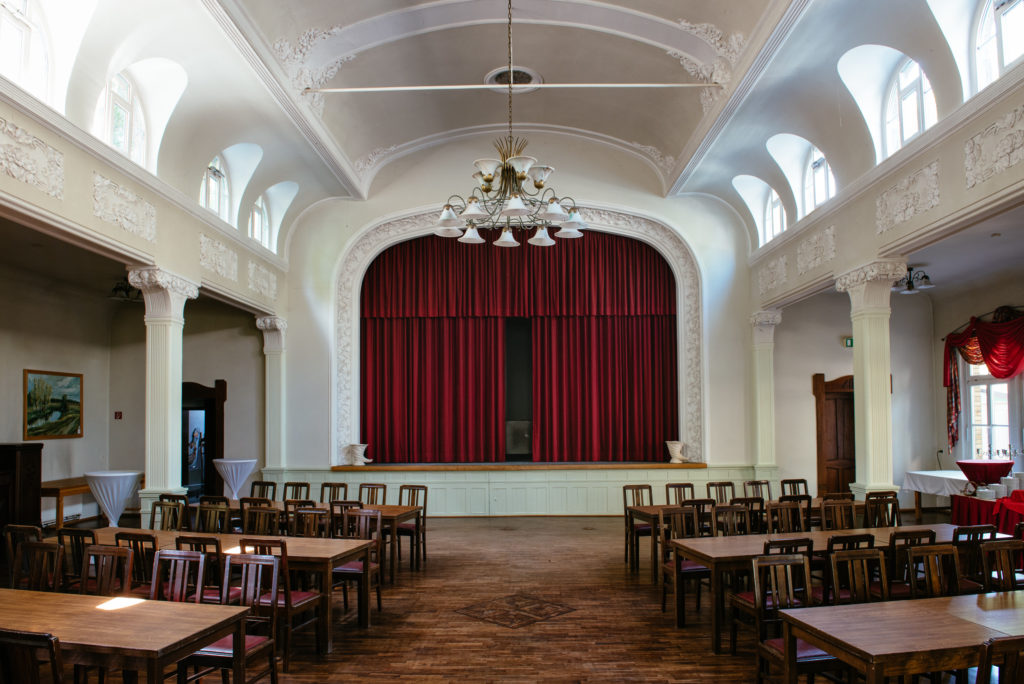 Ein großer Saal mit holzernen Tischen und Stühlen, einem roten Vorhang und weißen Wänden.