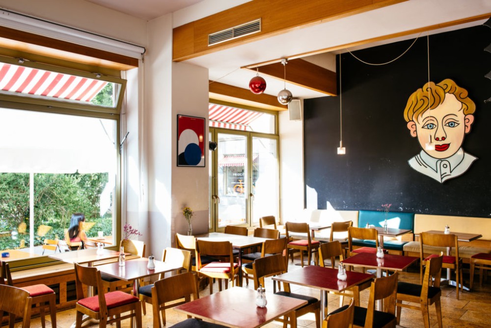 Helles Café mit bunten Stühlen, Holztischen und einer großen Wandzeichnung.